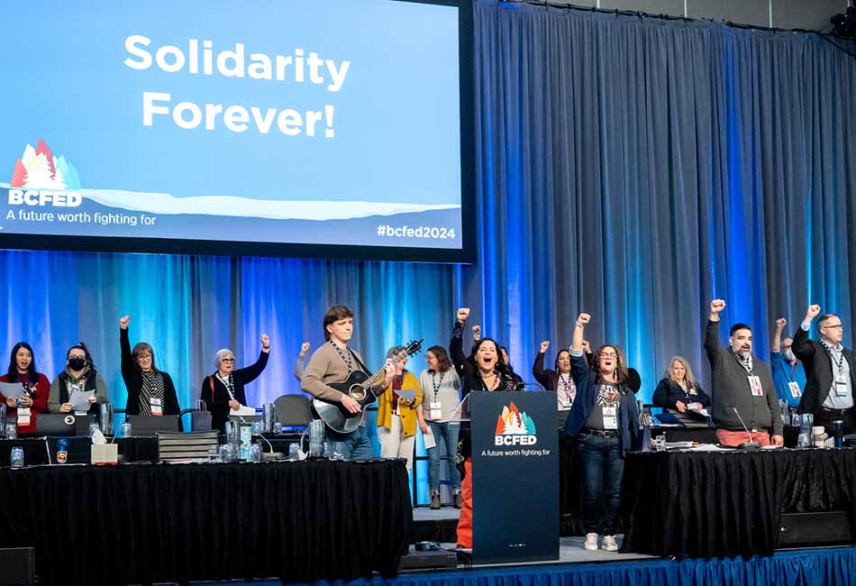 A convention stage with a slide reading "Solidarity forever!" and a lineup of over a dozen people singing with raised fists, with one person accompanying them on guitar