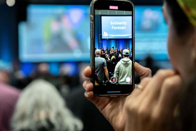 A delegate looks at their iPhone camera, which is displaying a stage where people are singing Solidarity Forever!