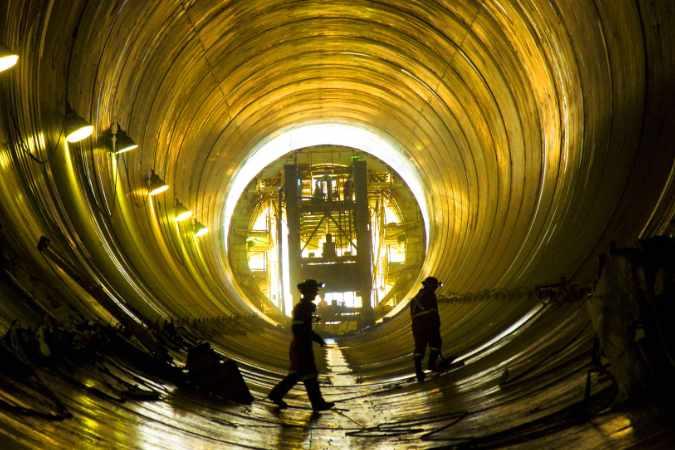 Two backlit construction workers walking through a gigantic conduit