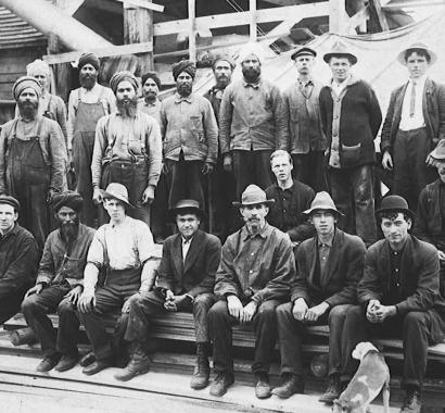 An old black-and-white photograph of about two dozen Punjabi and White workers posing outside a mill. There is a small dog in the foreground.